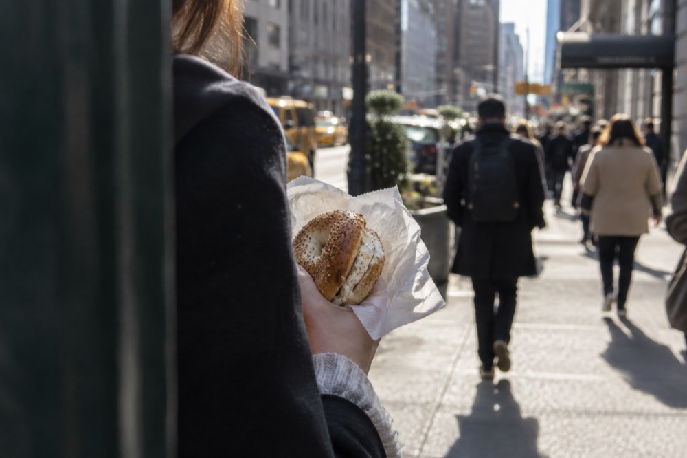 A person holding a toasted everything bagel with cream cheese wrapped in deli paper