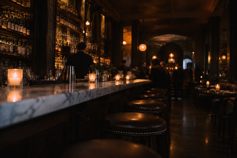 A candlelit speakeasy interior with a long marble bar and stools