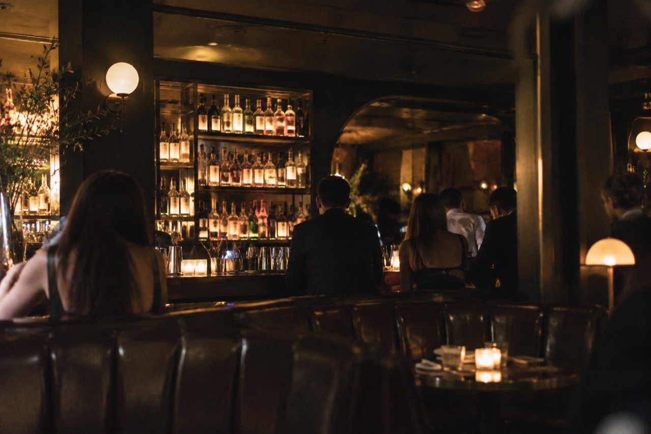 A dimly lit hidden bar room with leather booths and a backlit shelf of liquor bottles