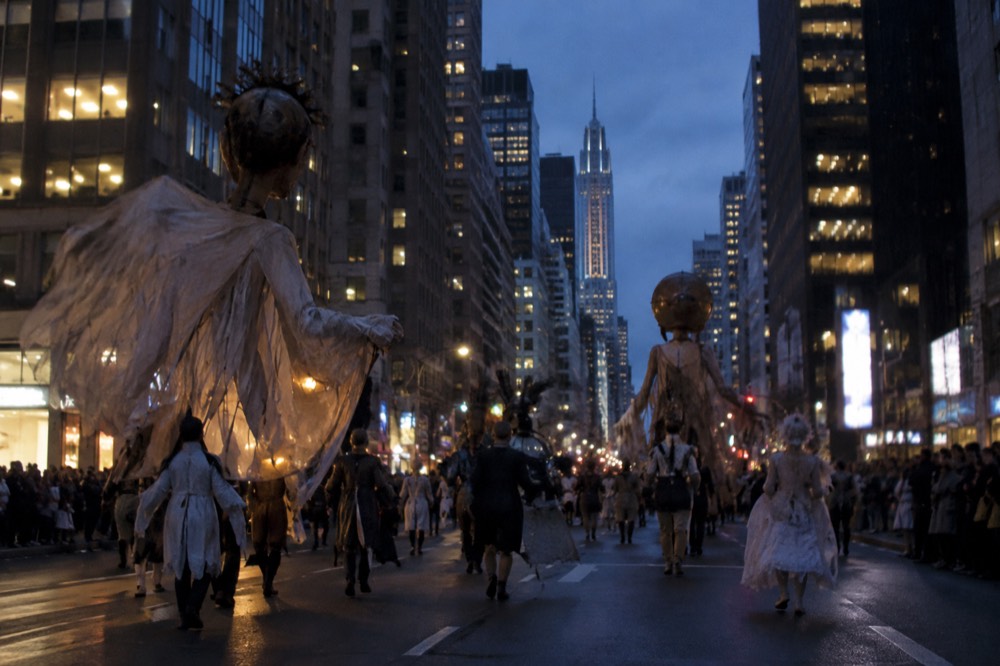 Giant puppets and costumed marchers moving along a wide avenue at night