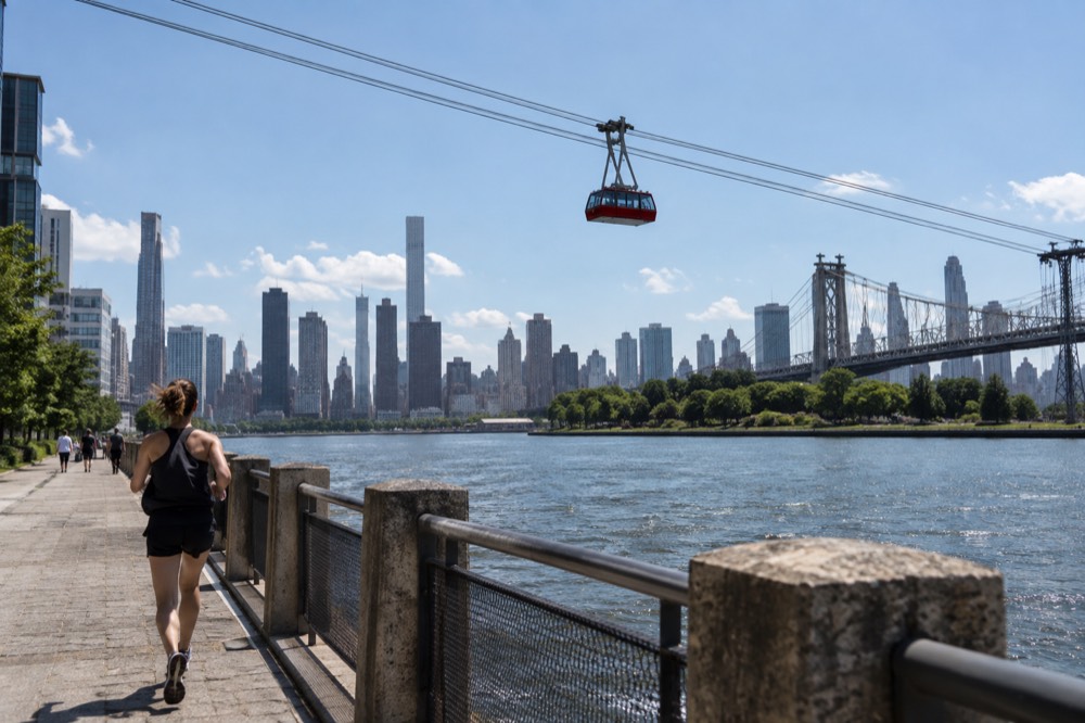 A cable car crossing high over the East River toward a low green island with the Manhattan skyline behind