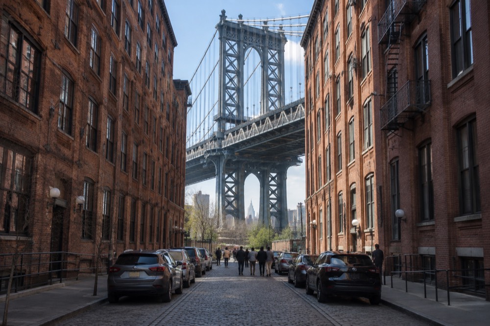 A cobblestone street in Brooklyn with the Manhattan Bridge framed between brick buildings overhead