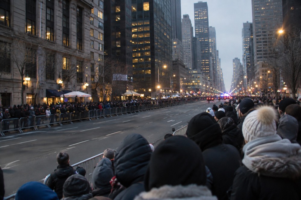 Spectators in winter coats line a barricaded Manhattan street before dawn waiting for the parade