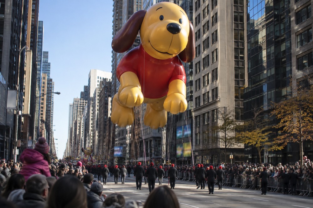 A giant helium balloon shaped like a cartoon character floats above a cheering crowd on a city avenue