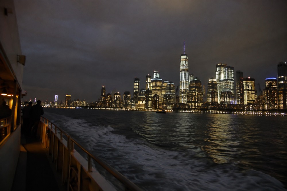 A boat cruising the harbor at night with the Manhattan skyline lit up behind it