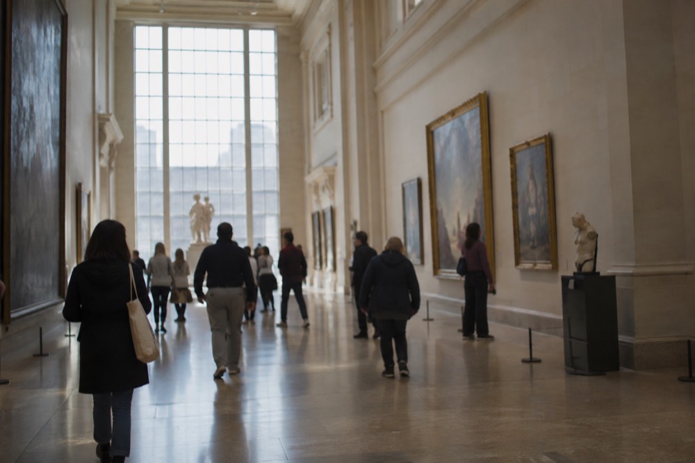 Visitors walking through a sunlit museum gallery with tall ceilings and paintings on the walls