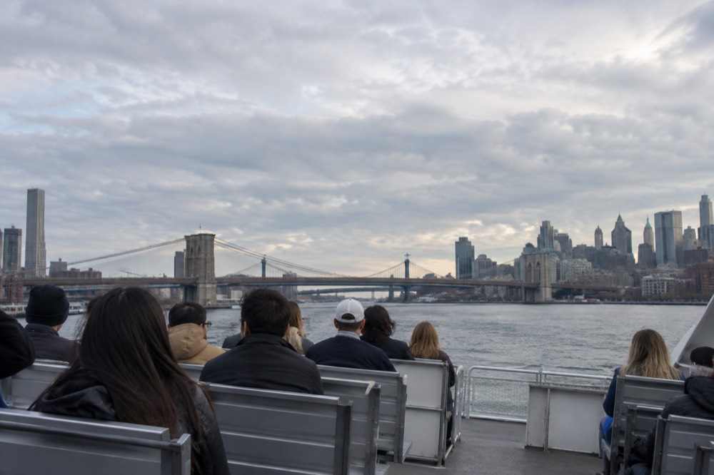 Passengers seated on the open upper deck of a ferry with the East River bridges in the distance
