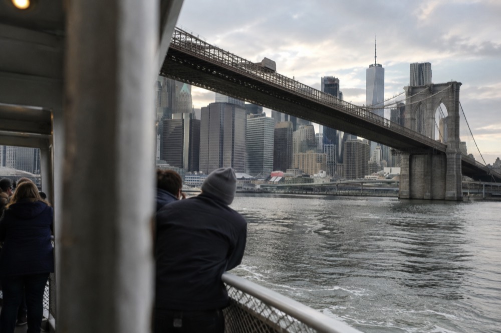 A ferry passing under the Brooklyn Bridge with passengers at the rail
