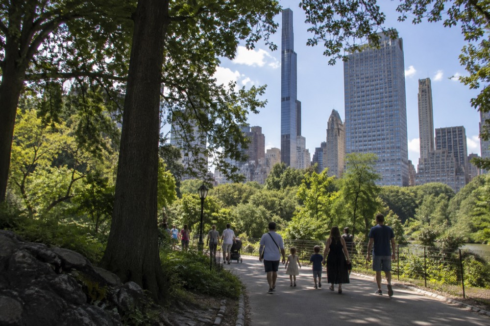 Families walking along a path beside Central Park with trees and skyline towers behind