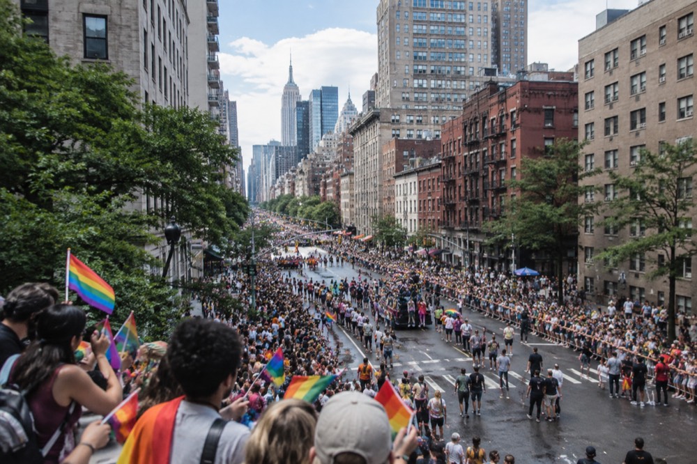 Crowds with rainbow flags line a wide Manhattan avenue under a bright sky