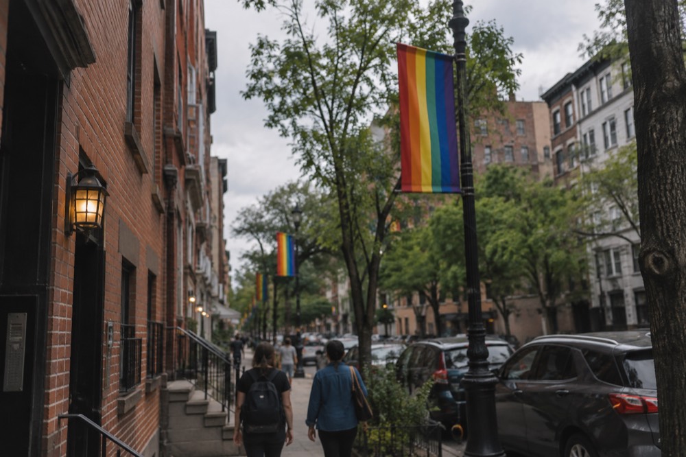 Rainbow flags hang from lampposts along a narrow Village street with old brick buildings