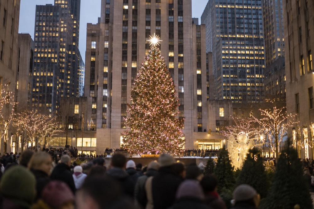 Crowds gathered around a lit Christmas tree in a plaza between tall buildings in the evening