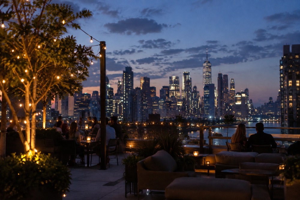 A rooftop bar at dusk with string lights, lounge seating and the Manhattan skyline glowing behind it