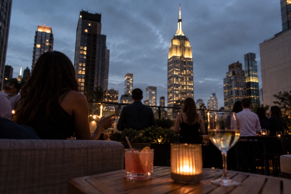 People with drinks at a rooftop bar looking toward the Empire State Building lit up at night