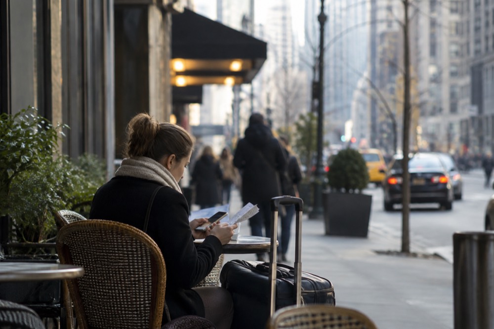 A traveler comparing paper tickets and a phone at a café table near a New York hotel
