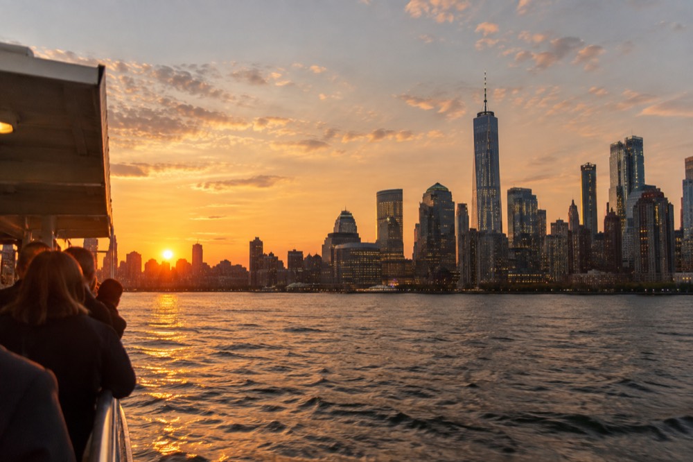 A sightseeing boat on the Hudson River at sunset with the illuminated Manhattan skyline behind it