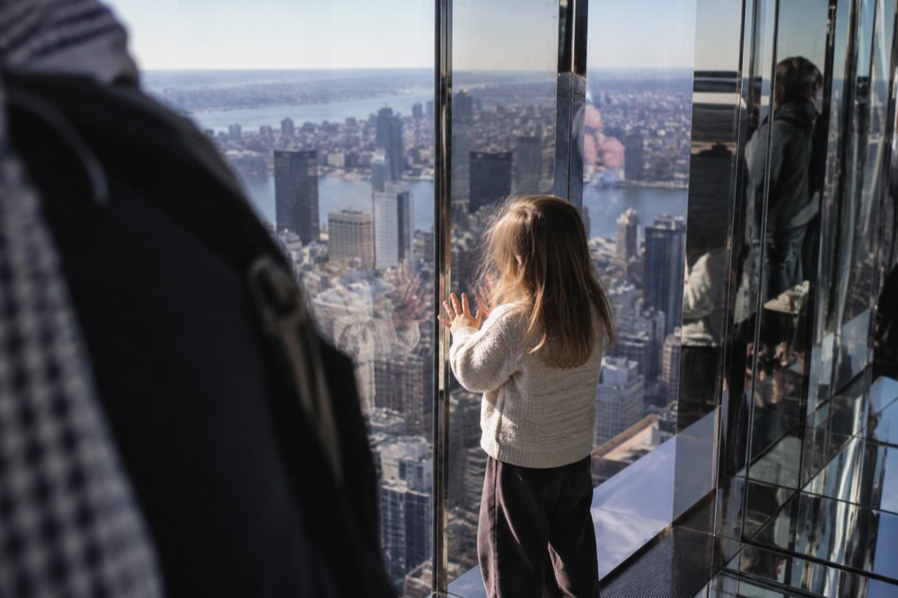 A child pressing hands against a large window in a mirrored observation room high above a city