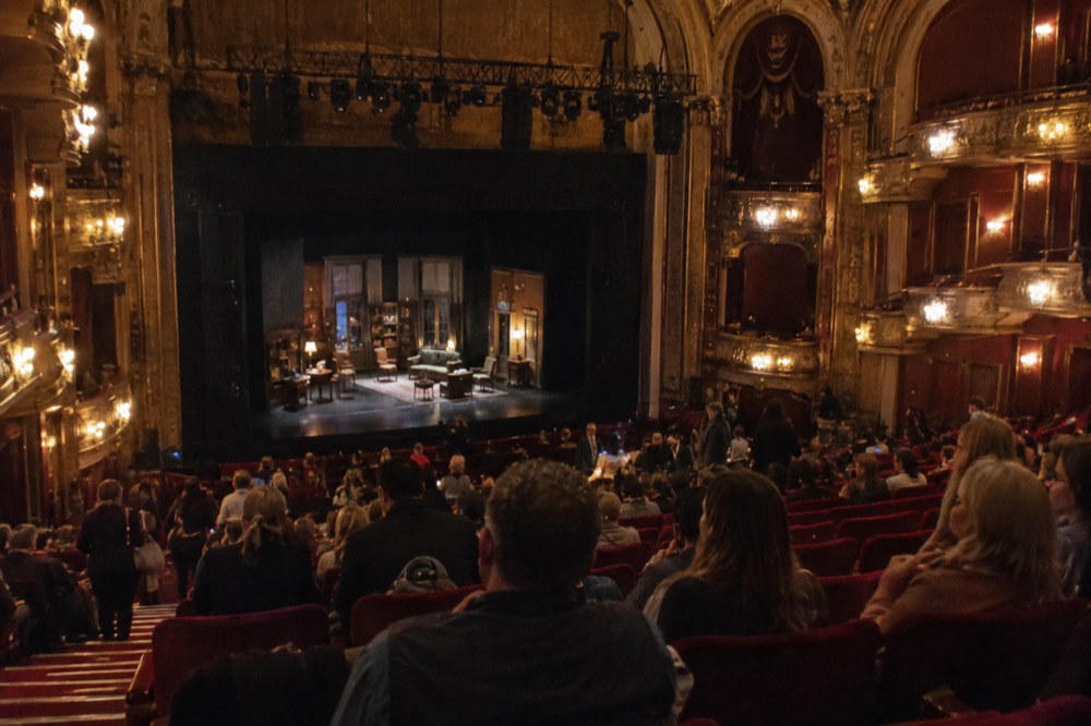 A theater interior with a lit stage and rows of red seats filling with an audience