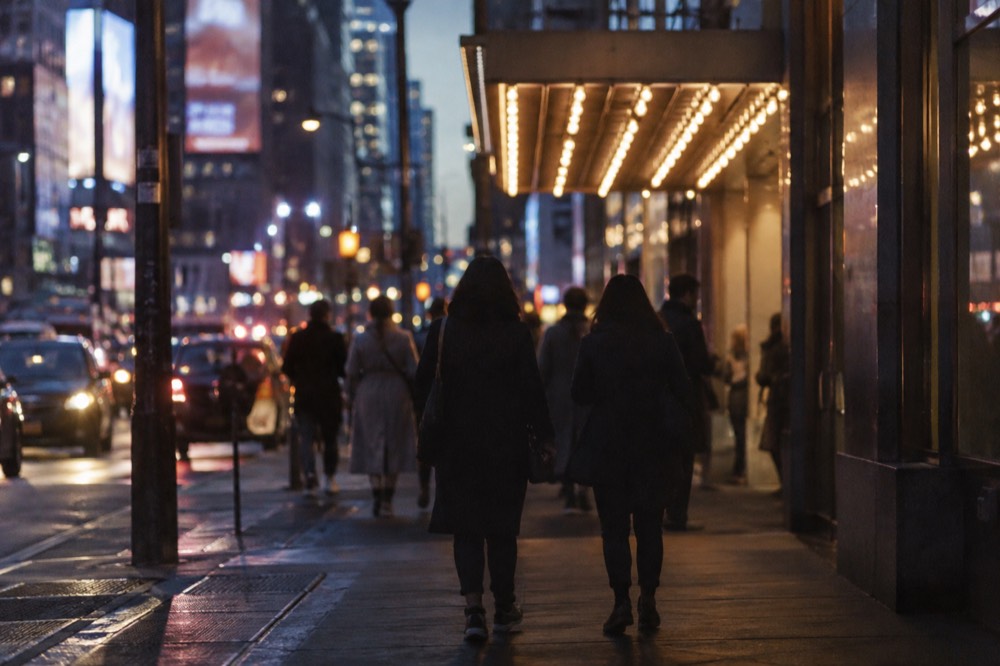 A Broadway sidewalk at night with a lit marquee and theatergoers heading toward the entrance