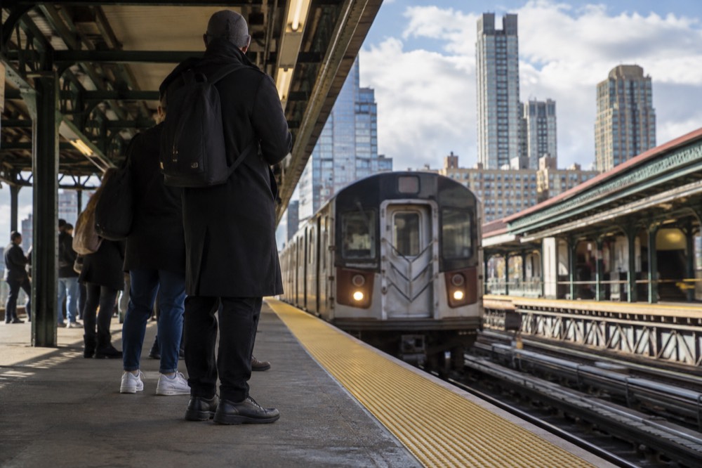 A subway platform in Manhattan with a train pulling in, passengers waiting behind the yellow safety line