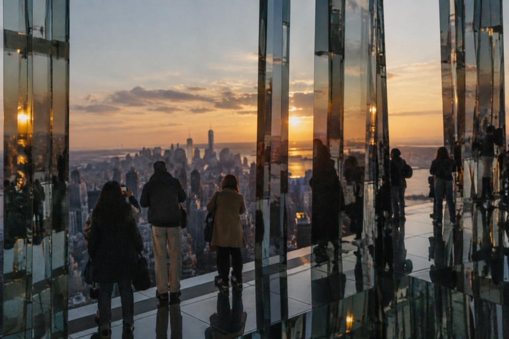 Visitors in a mirrored observation room at an observation deck at sunset, with the Manhattan skyline reflected in the glass