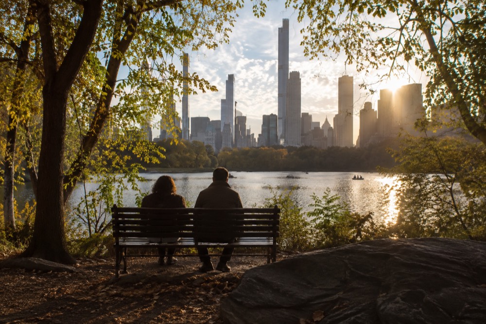 Two people sitting on a park bench overlooking a lake and city skyline through trees