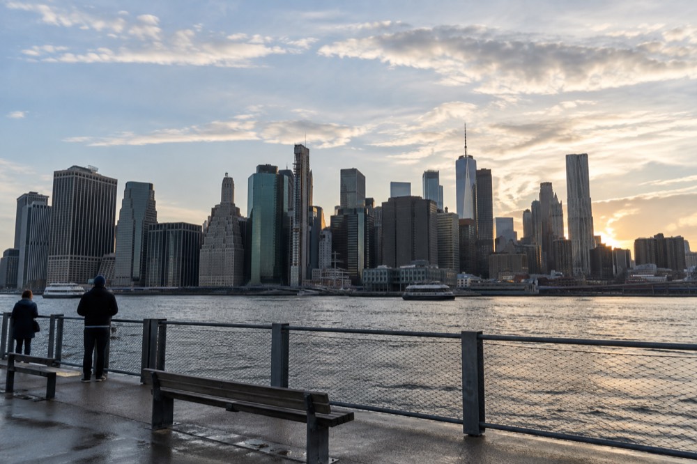 The Manhattan skyline seen across the East River from a waterfront promenade in late afternoon light