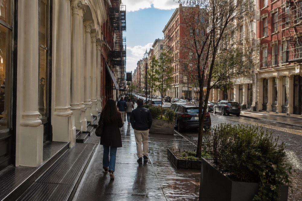 A narrow SoHo street with cast-iron building facades and shoppers walking in late morning sun