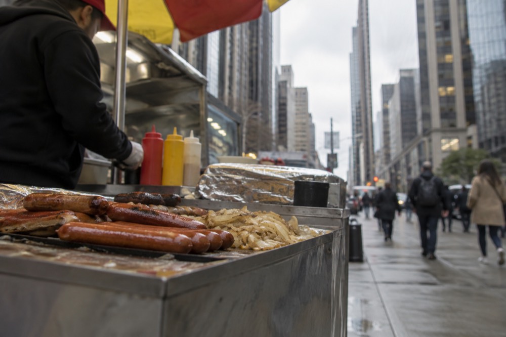 A hot dog cart on a Midtown sidewalk with office towers rising behind it