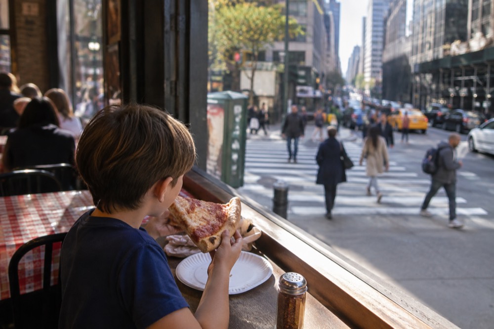 A child holding a large folded slice of pizza at a counter with checked tables