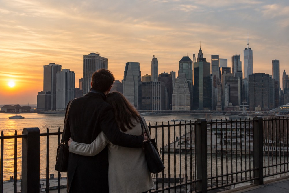 A couple standing on the Brooklyn Heights Promenade at sunset with the Lower Manhattan skyline behind them