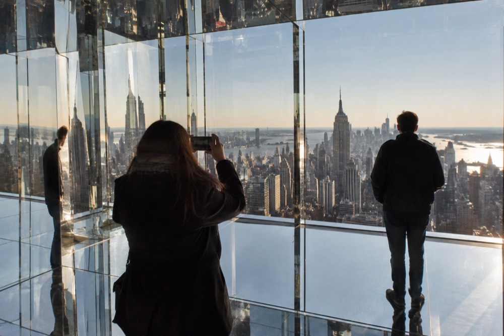 Two people in a mirrored room at an observation deck with the Manhattan skyline reflected around them